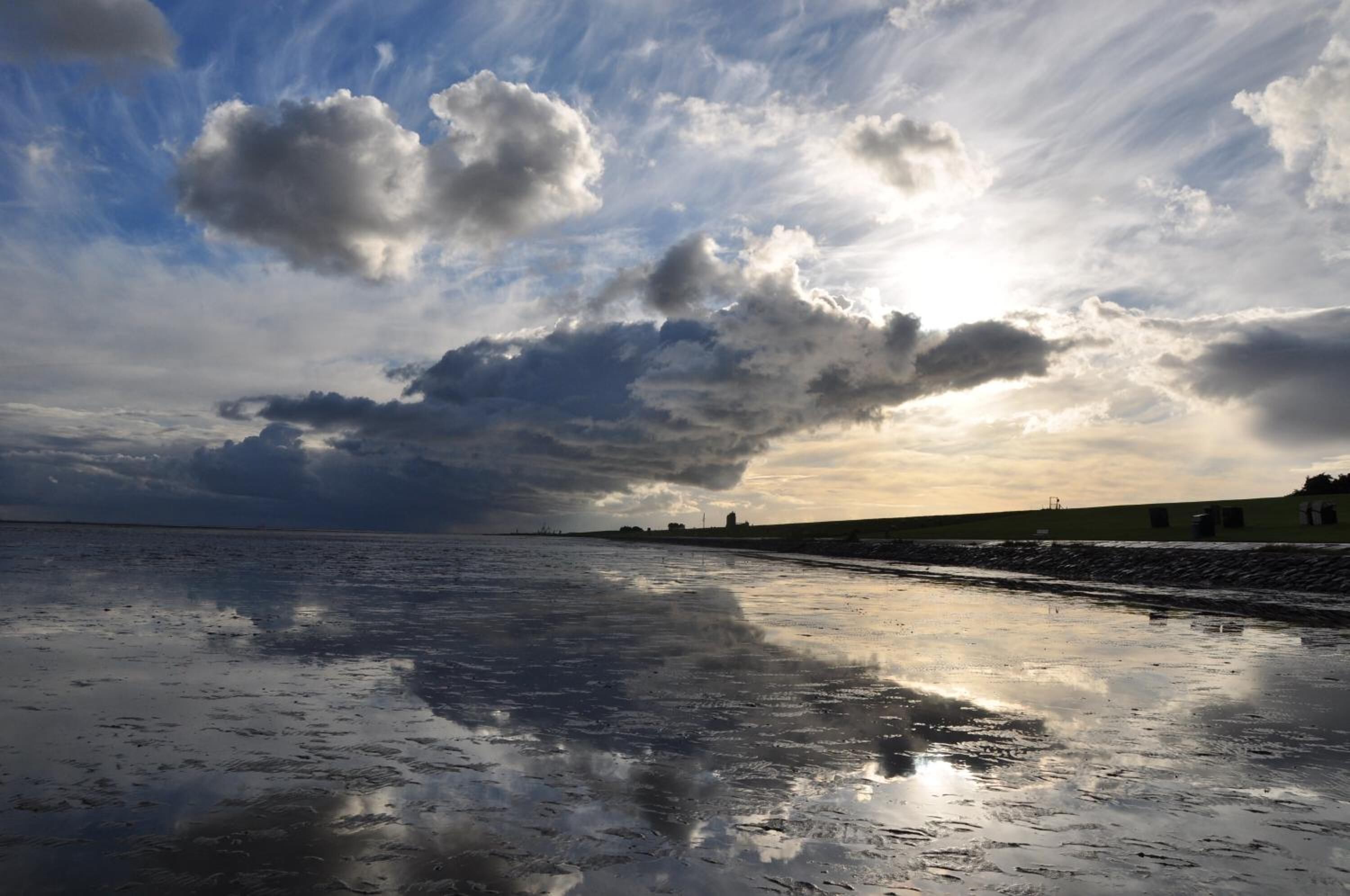 Een strand met een nat oppervlak dat wolken en een verre kustlijn weerspiegelt.