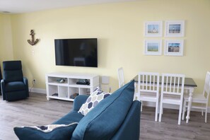 Un salón con un sofá color turquesa, un mueble de televisión blanco, una mesa de comedor con sillas blancas y un televisor montado en la pared.