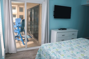 Un dormitorio con cama, cómoda, televisor en la pared y puerta corrediza de vidrio que da a un área exterior con sillas azules.