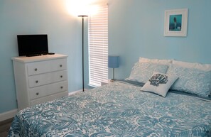 Ein Schlafzimmer mit einem Bett, einer Kommode, einem Fernseher auf der Kommode, einer Lampe und einem gerahmten Bild an der Wand.