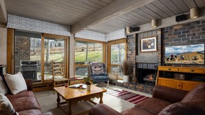 A cozy living room with a fireplace, leather sofas, and a large window overlooking a grassy area.