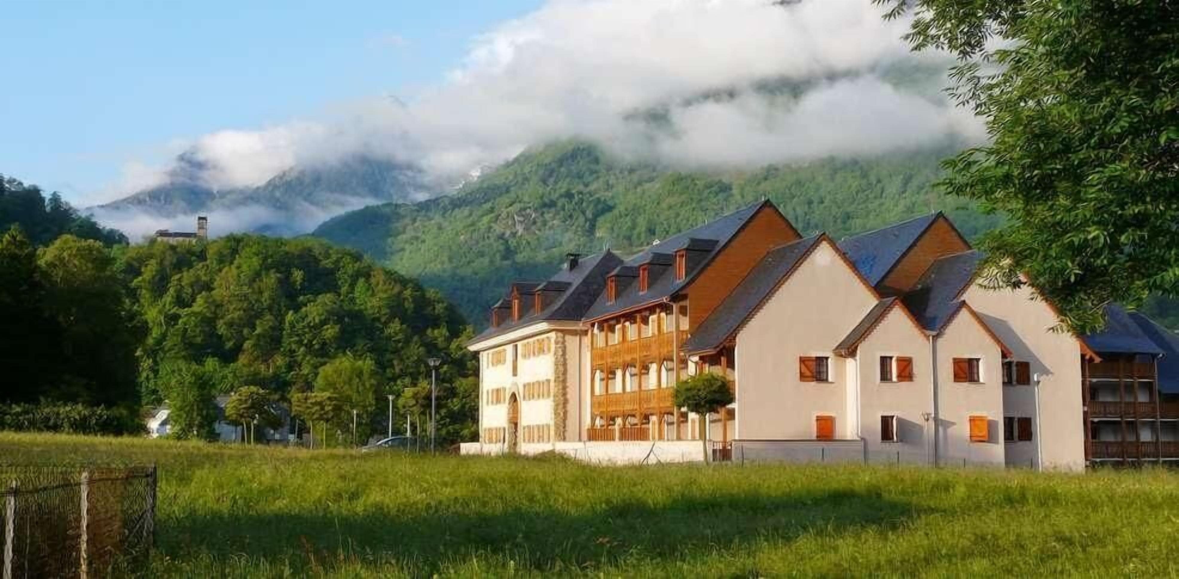 A row of traditional chalet-style buildings with sloped roofs, set against a backdrop of lush greenery and a mountain partially obscured by clouds.