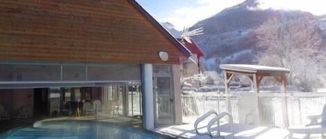 Una piscina con un edificio in legno sullo sfondo, una zona relax coperta e un paesaggio montuoso.