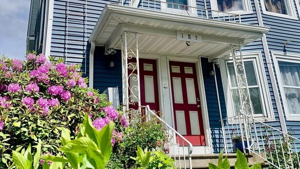 Una casa de dos pisos con una puerta roja, barandillas blancas y el número 185 en la fachada.