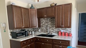 Una cocina con gabinetes marrón oscuro, salpicadero de azulejos, un lavamanos negro y una encimera con varios electrodomésticos y recipientes de almacenamiento.