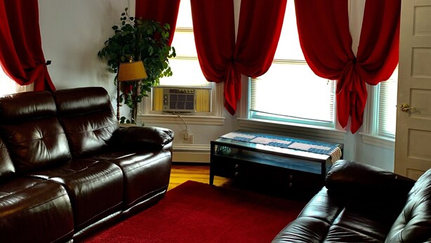 A living room with a ceiling fan, red curtains, a brown leather sofa, a coffee table, and a plant. - 2BR-Free Parking, Near Gillette & Providence-I-95 (Pawtucket)