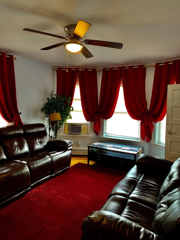 A living room with a ceiling fan, red curtains, a brown leather sofa, a coffee table, and a plant. - 2BR-Free Parking, Near Gillette & Providence-I-95 (Pawtucket)