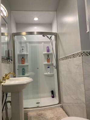 A bathroom with a glass-enclosed shower, a pedestal sink, and toiletries on shelves. - 2BR-Free Parking, Near Gillette & Providence-I-95 (Pawtucket)