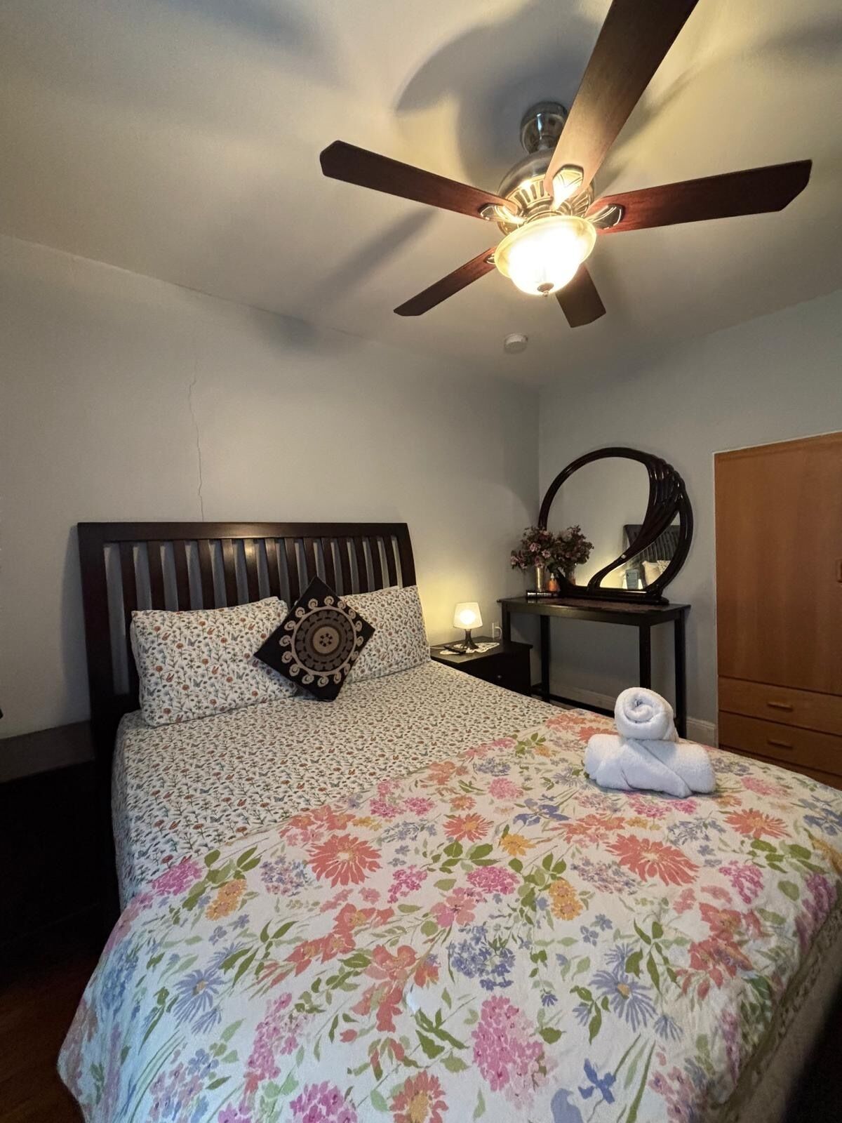 Une chambre à coucher comprenant un lit, un ventilateur de plafond, une table de chevet, un miroir et une armoire en bois.