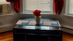 Una habitación con ventana, cortinas rojas, un florero con flores y una mesa con superficie de vidrio.