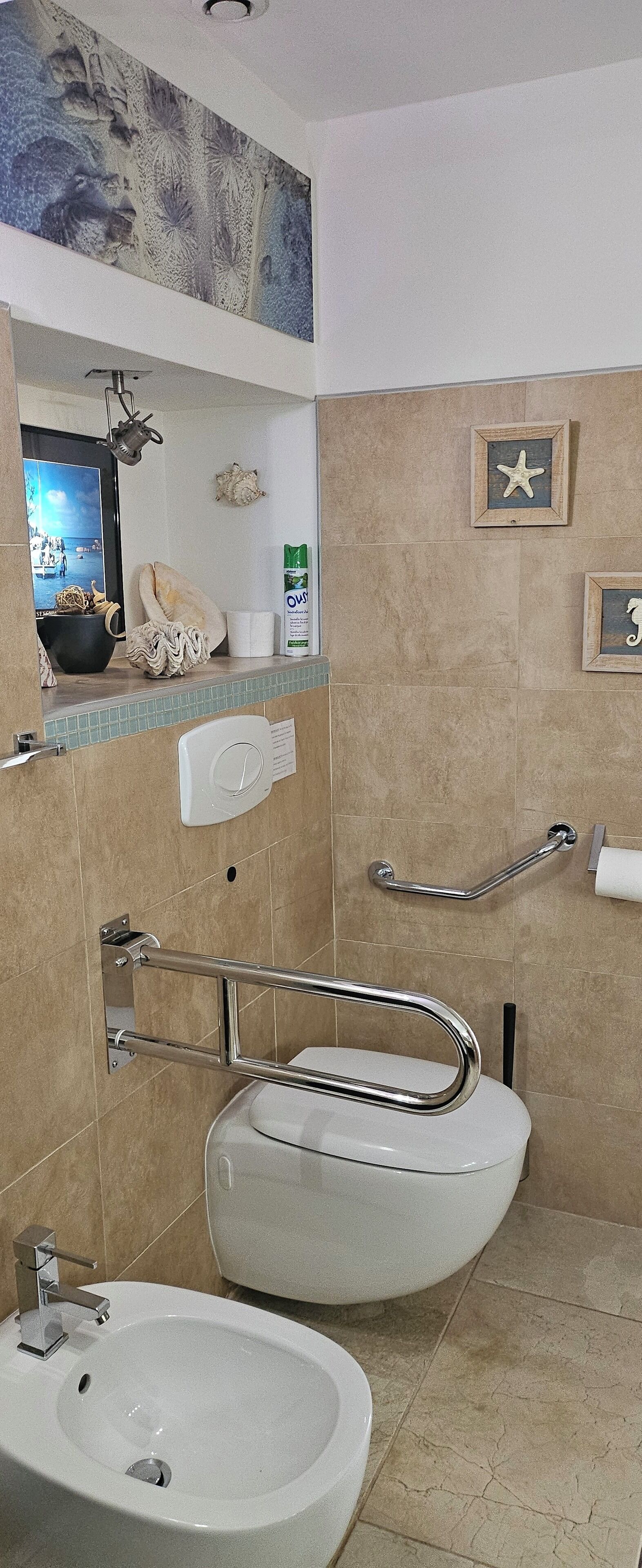 A bathroom with a white sink, a toilet, and a bidet. There is a mirror above the sink, and a shelf with decorative items.