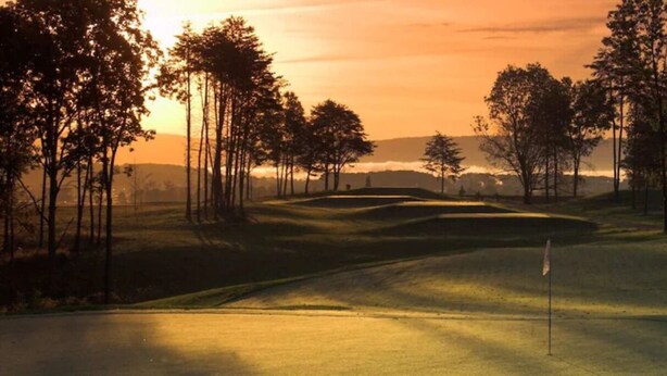 A golf course with a flagstick, surrounded by trees and rolling hills.