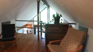 A cozy attic room with wooden flooring, a cushioned chair, a small table, and a staircase leading to an upper level.