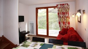 Una habitación con un sofá rojo, una mesa de comedor con mantel individual verde, un televisor sobre un mueble bajo y un ventanal corredizo que da a un jardín.