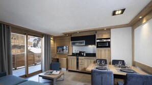 Una sala de estar moderna y bien iluminada, con un ventanal grande, una zona de comedor con una mesa para dos personas y una cocina con armarios de madera y electrodomésticos modernos.