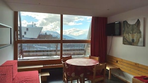 A room with a large window offering a mountain view, a round table with chairs, a red sofa, a wooden bench, and a mounted TV. - 50m from ski lifts, 4th floor, view mountain, balcony, tv, 30m², Les Arcs (Les Arcs)