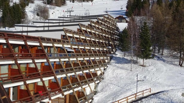 Een meerlaags hotel met een houten gevel en balkons, gelegen in een besneewd bergachtig gebied met skipistes op de achtergrond.