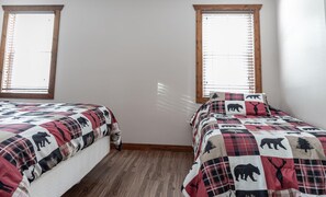 A room with two beds, each with a quilt featuring animal prints. There are two windows with wooden frames and blinds.