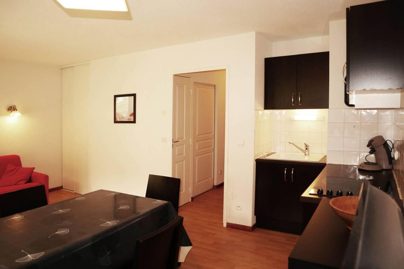 A compact kitchen with a dining area, featuring a black table with leaf patterns, a red sofa, and a white door leading to another room.