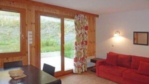 A room with a wooden floor, a red sofa, a dining table with chairs, and a large window with a view of greenery. - Ski-in/ski-out & foot of the slopes, 1st floor, view swimming pool, swimming pool, sauna, hammam (Les Orres)