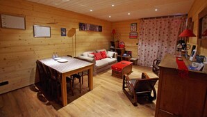 Une pièce confortable, aménagée dans le style d’un chalet, avec un intérieur en bois, une table à manger avec des chaises, un canapé avec des coussins rouges, un petit bureau en bois et une bibliothèque.