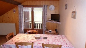 Ein gemütlicher Raum mit Holzvertäfelung, einem Esstisch mit gemustertem Tischläufer, zwei Holzstühlen, einem Fernseher auf einem Regal und einer Uhr an der Wand.