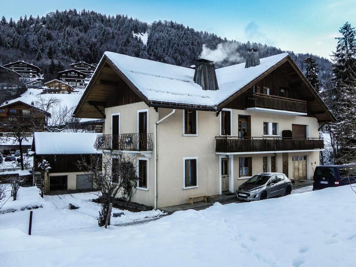 A two-story chalet with a balcony, surrounded by snow-covered trees and a mountainous backdrop.