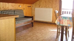 Ein gemütliches Blockhäuschen-Interieur mit Holzbalkendecke, einem karierten Sofa, einem Esstisch mit bunten Tischdecken und einer Heizkörper.