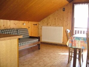 Ein gemütliches Blockhäuschen-Interieur mit Holzbalkendecke, einem karierten Sofa, einem Esstisch mit bunten Tischdecken und einer Heizkörper.