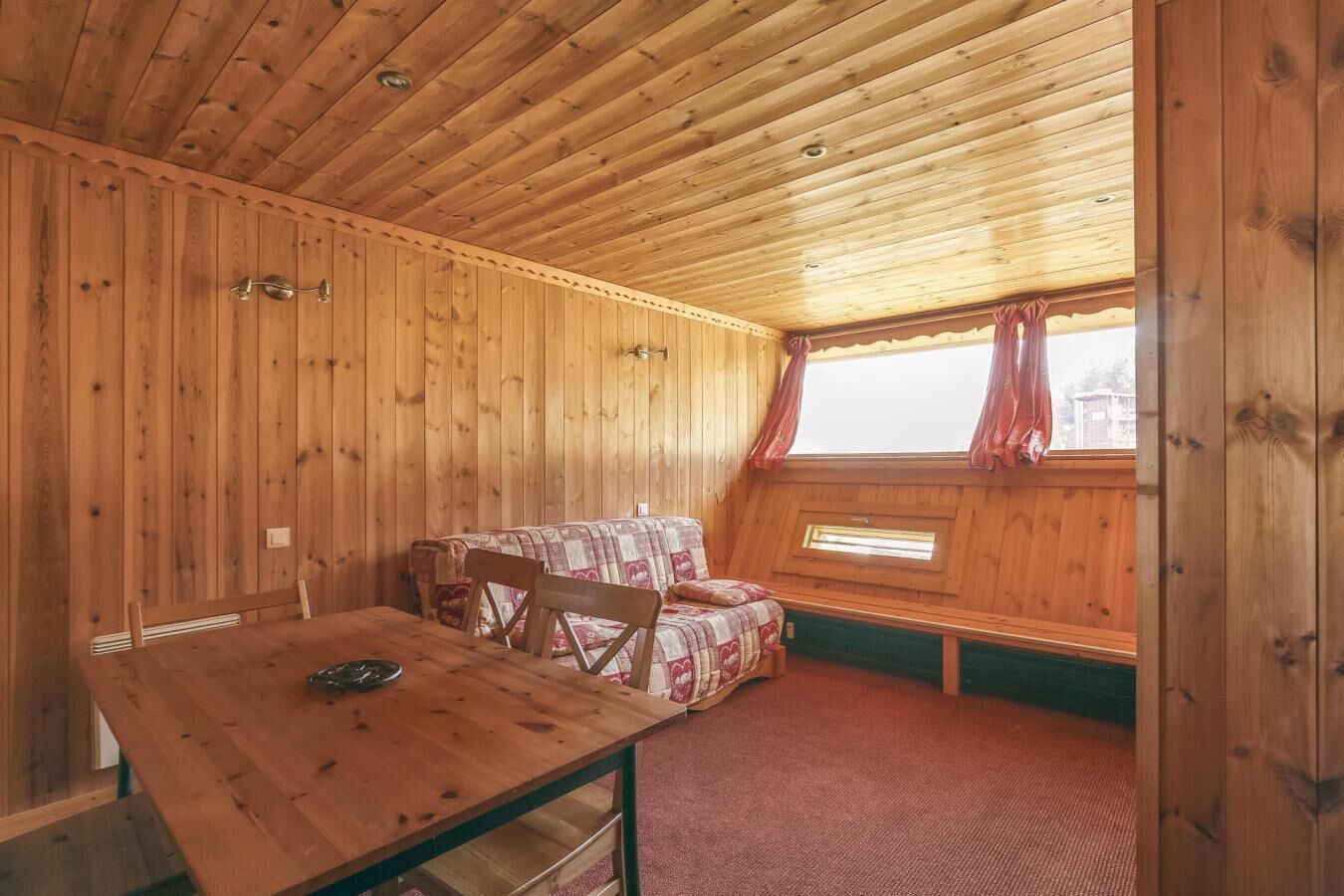 Una cabana de fusta amb una taula de dinar i dues cadires, una finestra amb cortines, i un sostre inclinat.