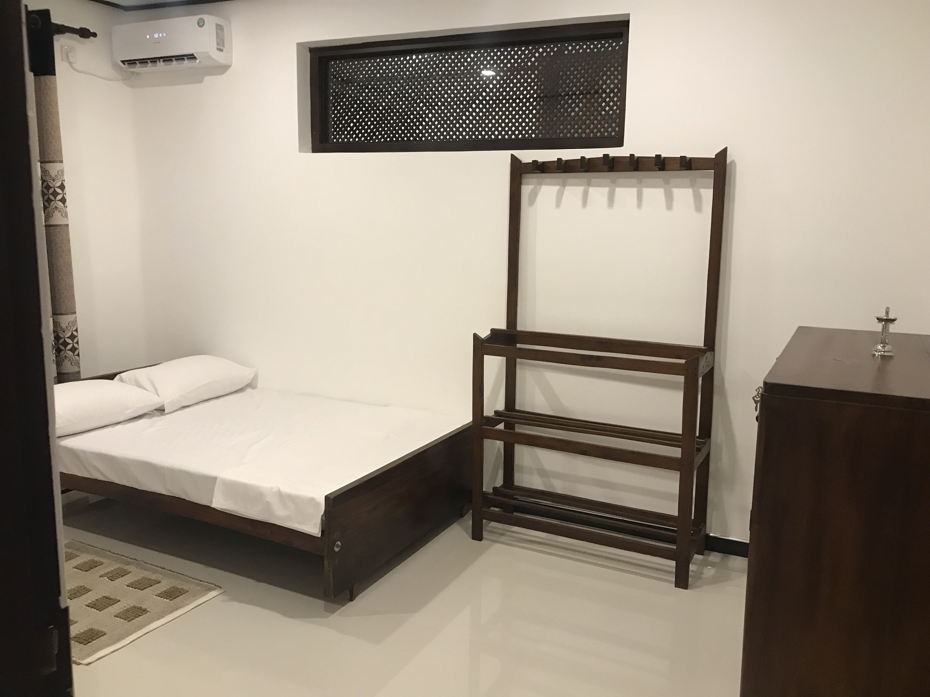 Un dormitorio con cama, un armario de madera, una ventana con diseño de celosía y un equipo de aire acondicionado.