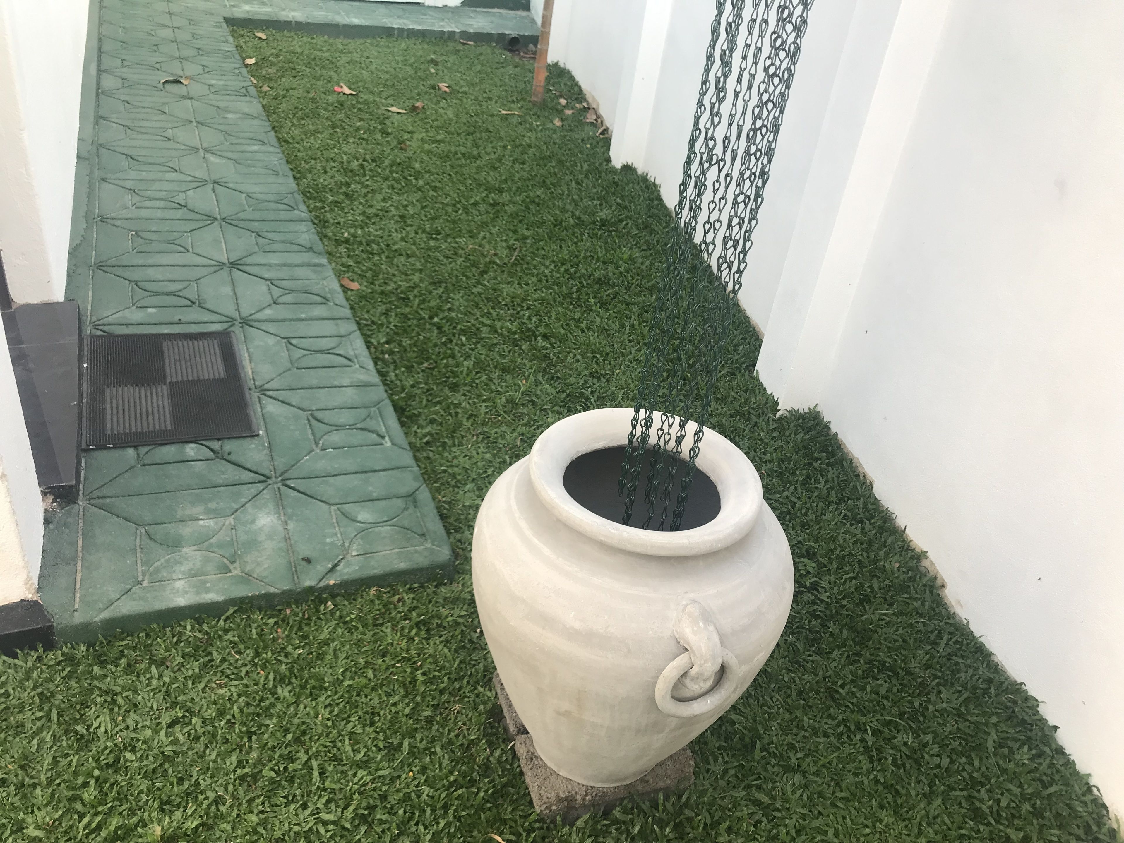 Um vaso branco com grade de metal, colocado sobre um tapete de grama artificial, ao lado de uma parede branca e um caminho com padrão verde.