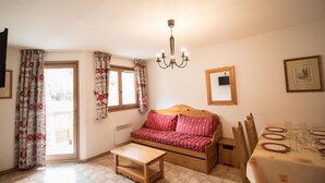 A cozy room with a wooden sofa, a small wooden table, a dining table set with plates and glasses, a chandelier, and a window with curtains. - 10m from ski slopes, 1st floor, balcony, tv, ski locker, 40m², Val Cenis (Lanslevillard)
