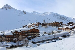 A snow-covered mountain resort with wooden buildings, a ski lift, and a road with parked cars.