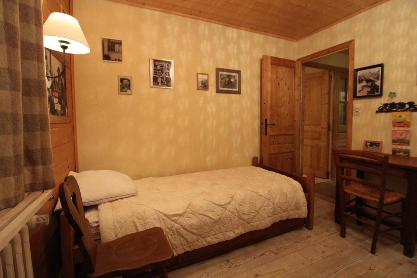 A small, cozy room with a single bed, a wooden desk, and a chair. There are several framed pictures on the wall.