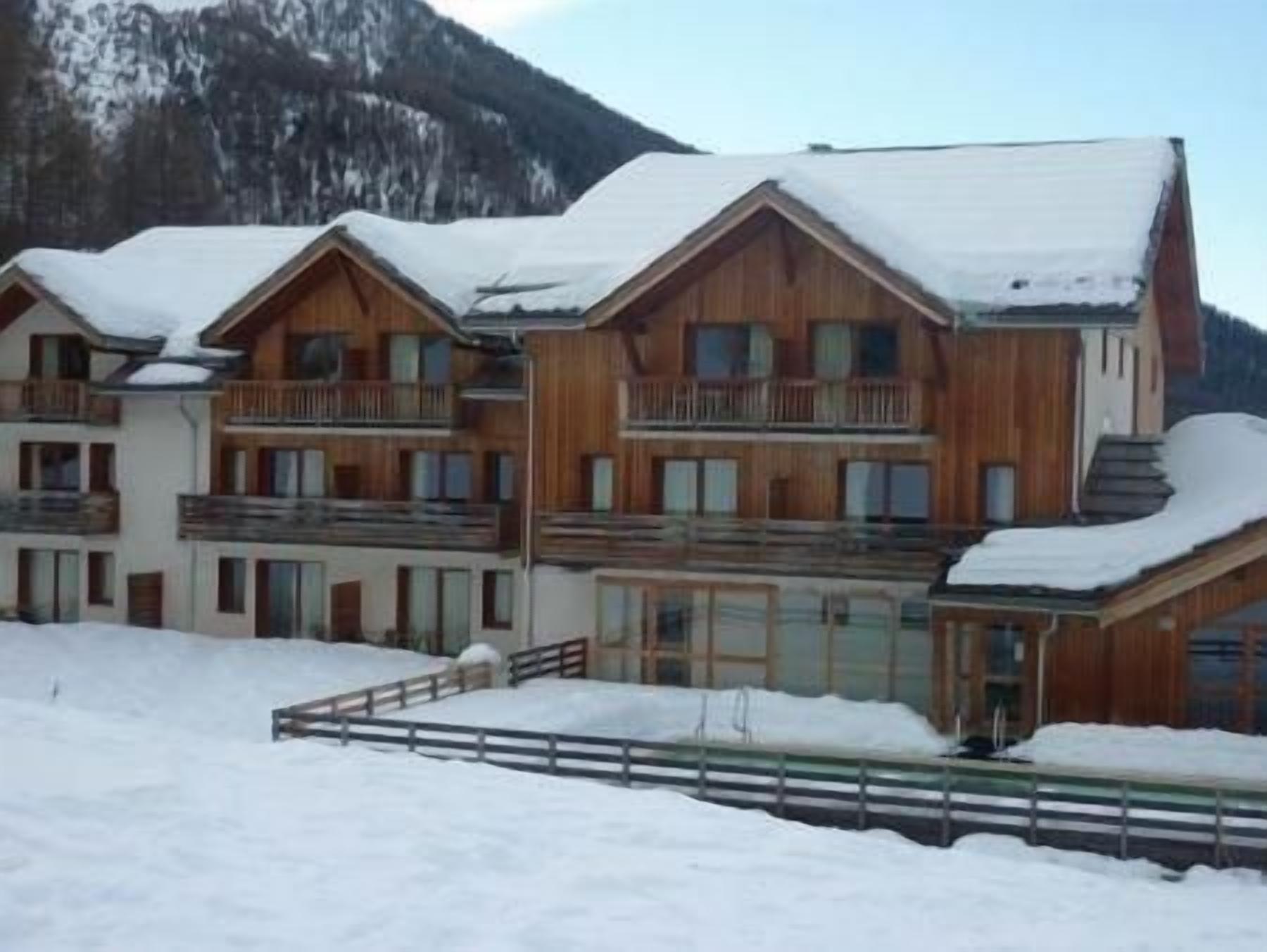 A multi-story wooden building with balconies, surrounded by snow-covered ground and mountains in the background.