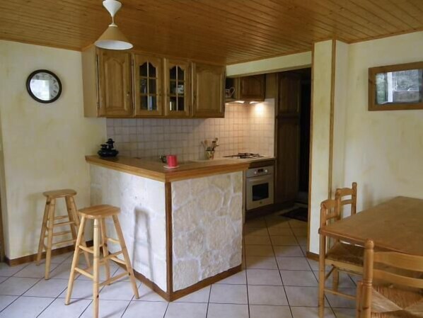 Une cuisine avec des meubles de rangement en bois, un îlot central doté de tabourets, une horloge au mur et une fenêtre.