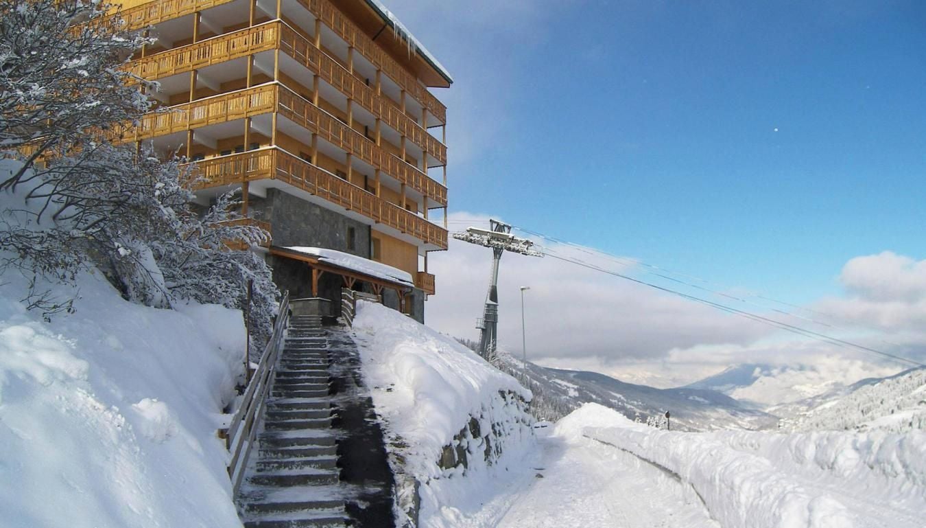 Un immeuble à plusieurs étages doté de balcons, un escalier menant à l’entrée et un paysage enneigé en arrière-plan.
