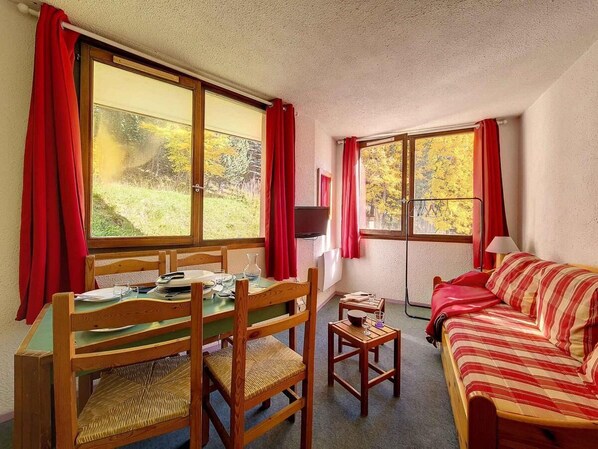 Ein Zimmer mit Esstisch, Stühlen, einem Bett und zwei Fenstern mit roten Vorhängen.