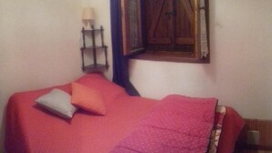Une chambre à coucher avec un lit, des oreillers, une table de chevet, une lampe et une fenêtre avec un volet en bois.