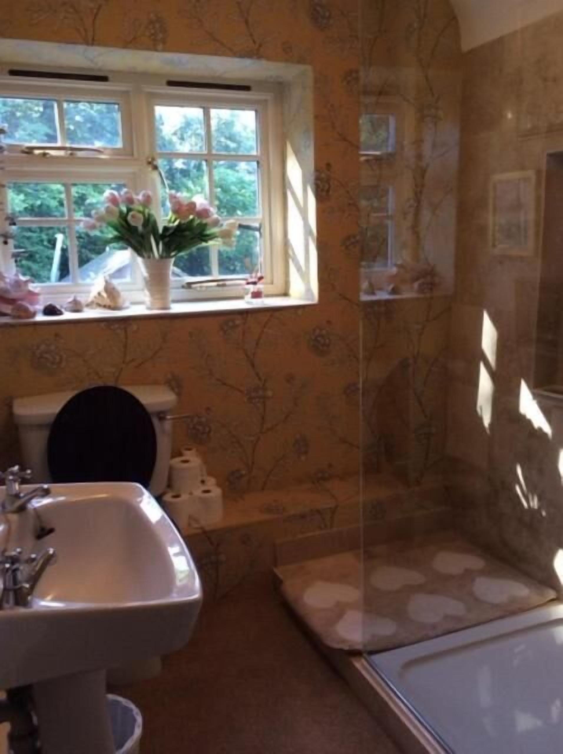 A bathroom with a sink, toilet, and shower. There is a window with flowers on the windowsill.