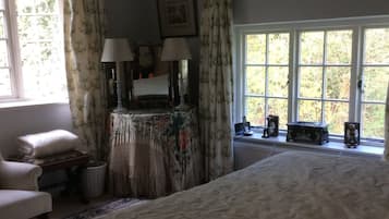A bedroom with a large bed, a chair, a vanity with a mirror, and a window with curtains.