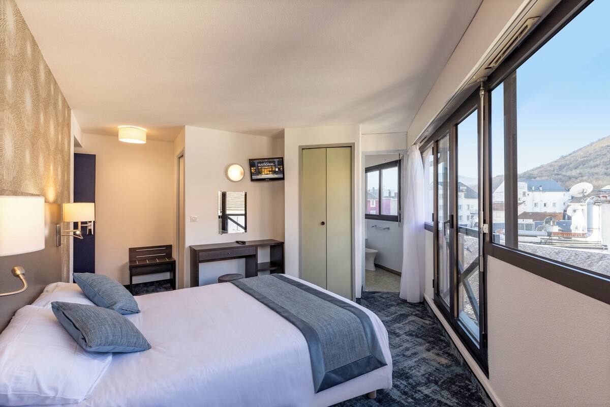 Habitación de hotel con cama, escritorio, espejo y vistas al exterior.