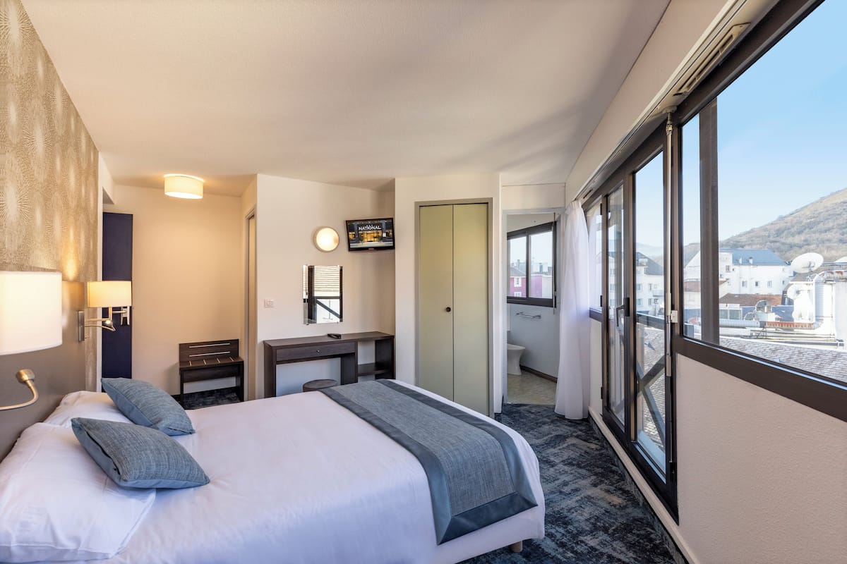 Quarto de hotel com cama, escrivaninha, espelho, televisão e vista para o exterior.
