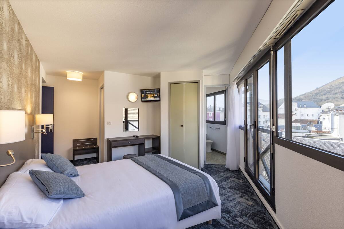 Habitación de hotel con cama, escritorio, espejo y vistas al exterior.