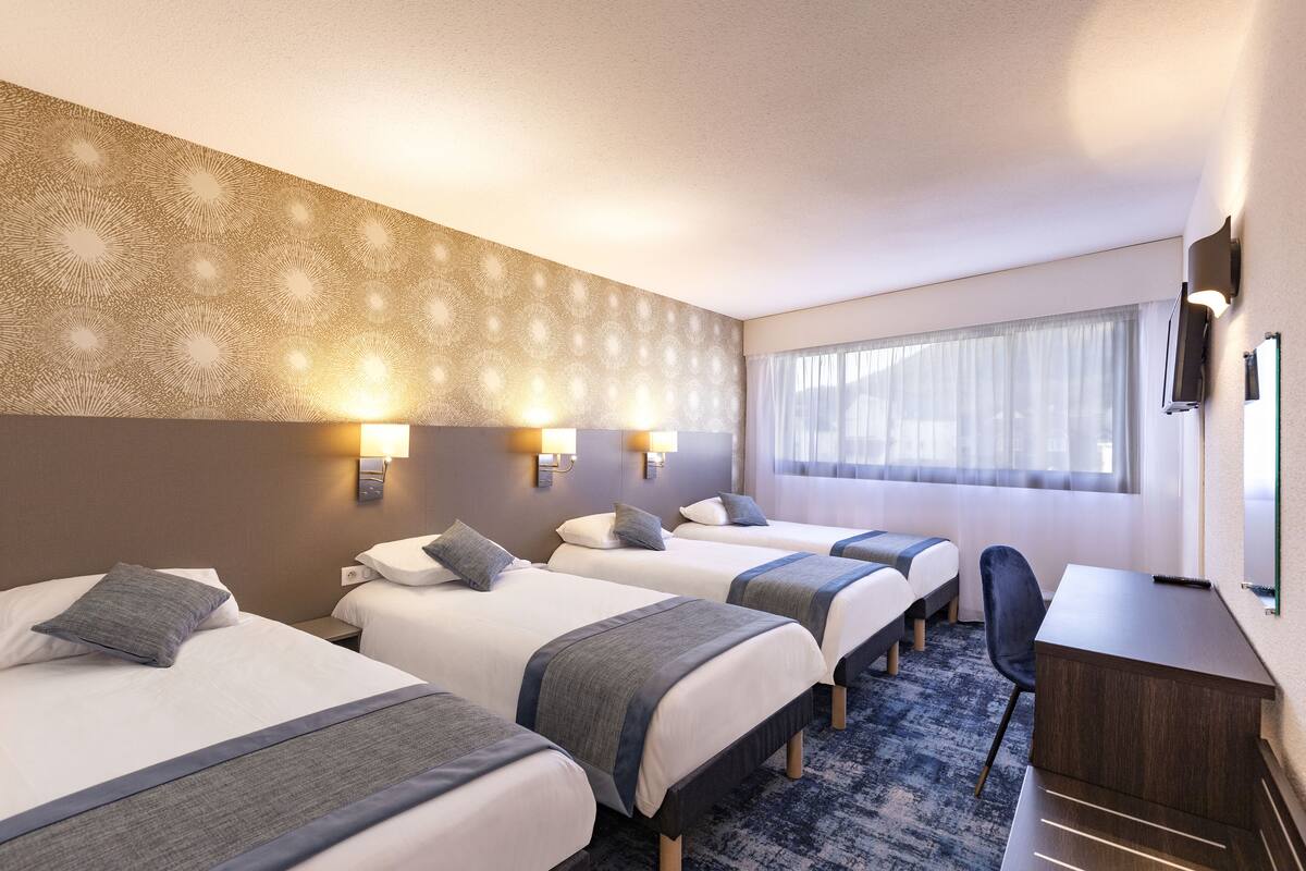 Habitación de hotel con tres camas, un escritorio y un ventanal con cortinas.