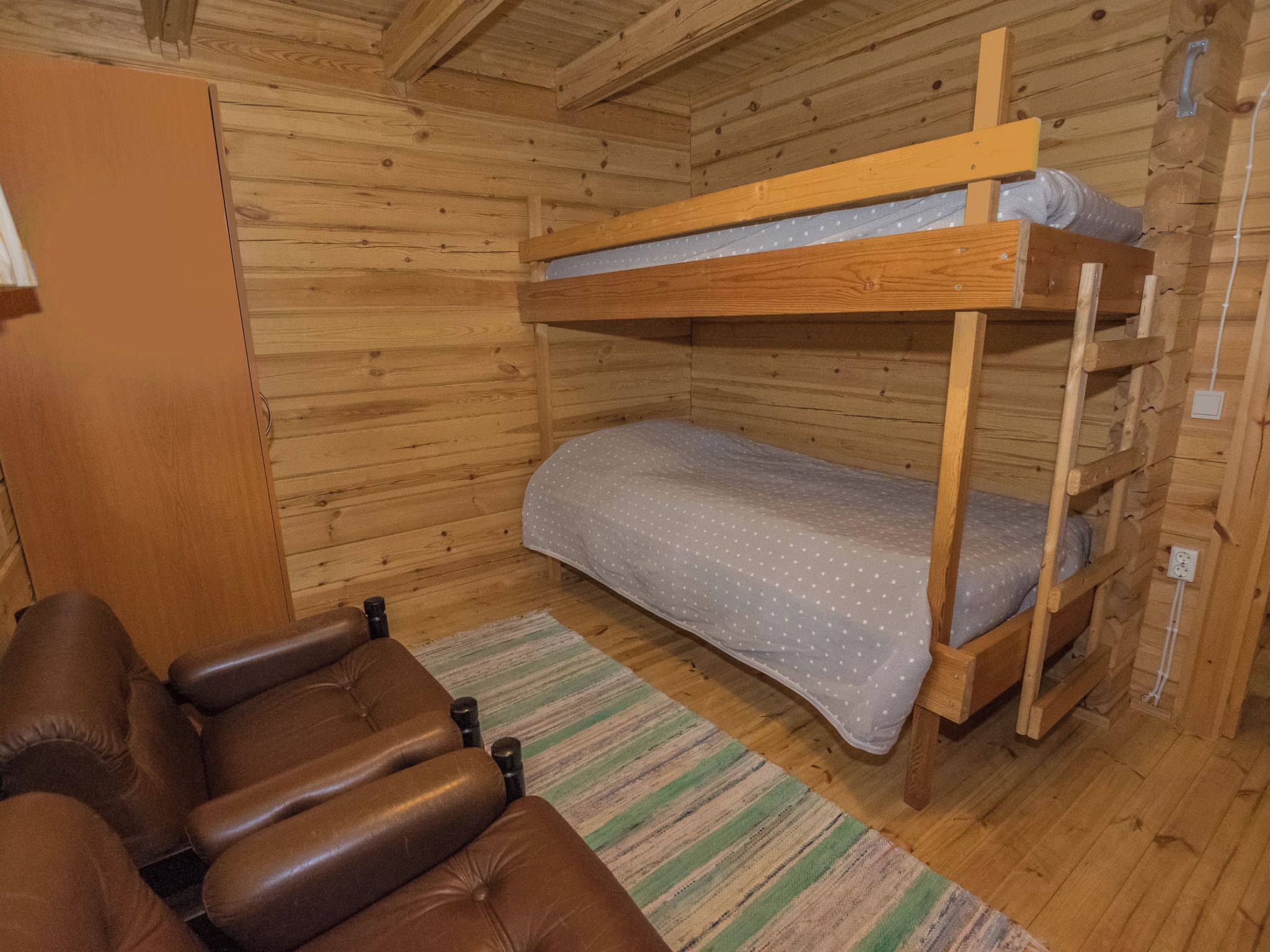 A bunk bed with a mattress and a wooden ladder, a brown leather chair, and a striped rug on a wooden floor.