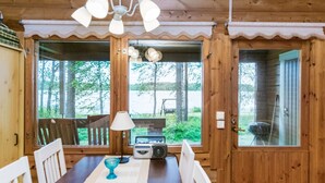 Interior de una cabaña de madera con una mesa de comedor, sillas, una radio y ventanas amplias que ofrecen vistas del exterior.