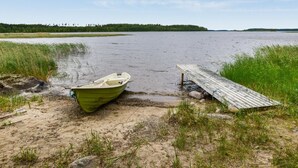 Un bote atracado en una orilla arenosa con un muelle de madera que se adentra en un lago tranquilo, rodeado de zonas verdes y una orilla boscosa.