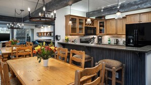 A modern kitchen with wooden cabinets, a central island with bar stools, pendant lighting, and a dining area with a wooden table and chairs. - Top of the Village~Ski-in/out~HT~Pool~Grill~Parking (Snowmass Village)